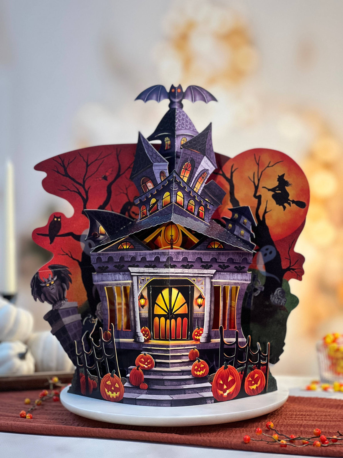 A 3D papercraft Halloween haunted house centerpiece on a white plate, featuring a purple gothic house with glowing windows, jack-o'-lanterns, a witch on a broomstick against a full moon, and spooky trees, set against a warm, softly blurred background.