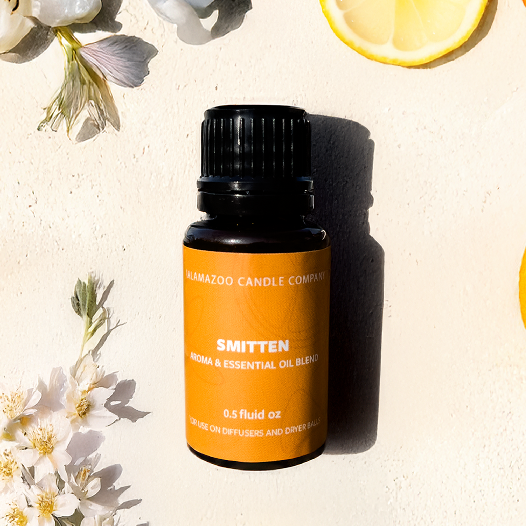 A  bottle of Kalamazoo Candle Company 'Smitten' essential oil blend, styled in a flat lay with lemon slices, white flowers, and greenery on a textured cream background.