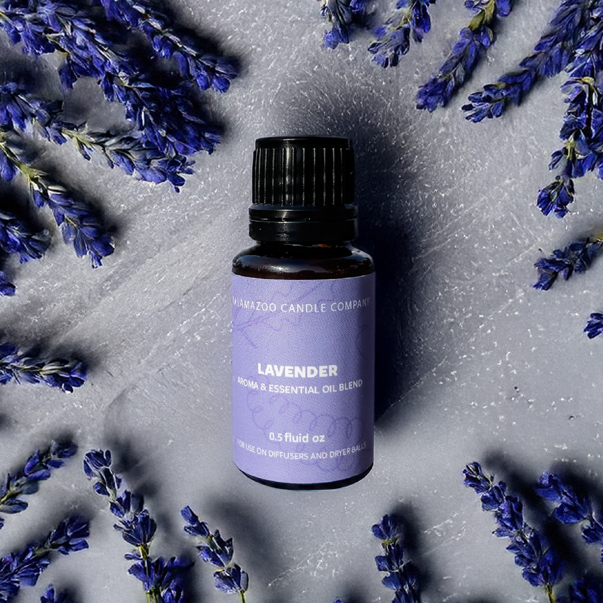 A small amber glass bottle of lavender essential oil blend with a black cap and a lavender-purple label, centered on a textured gray surface and surrounded by scattered purple lavender sprigs.