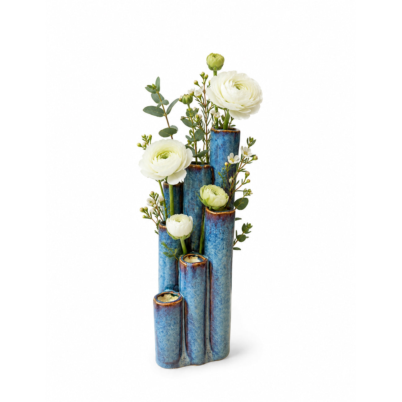 A modern multi-tier ceramic vase in a textured blue glaze, styled with white ranunculus and green foliage, set against a plain white background.