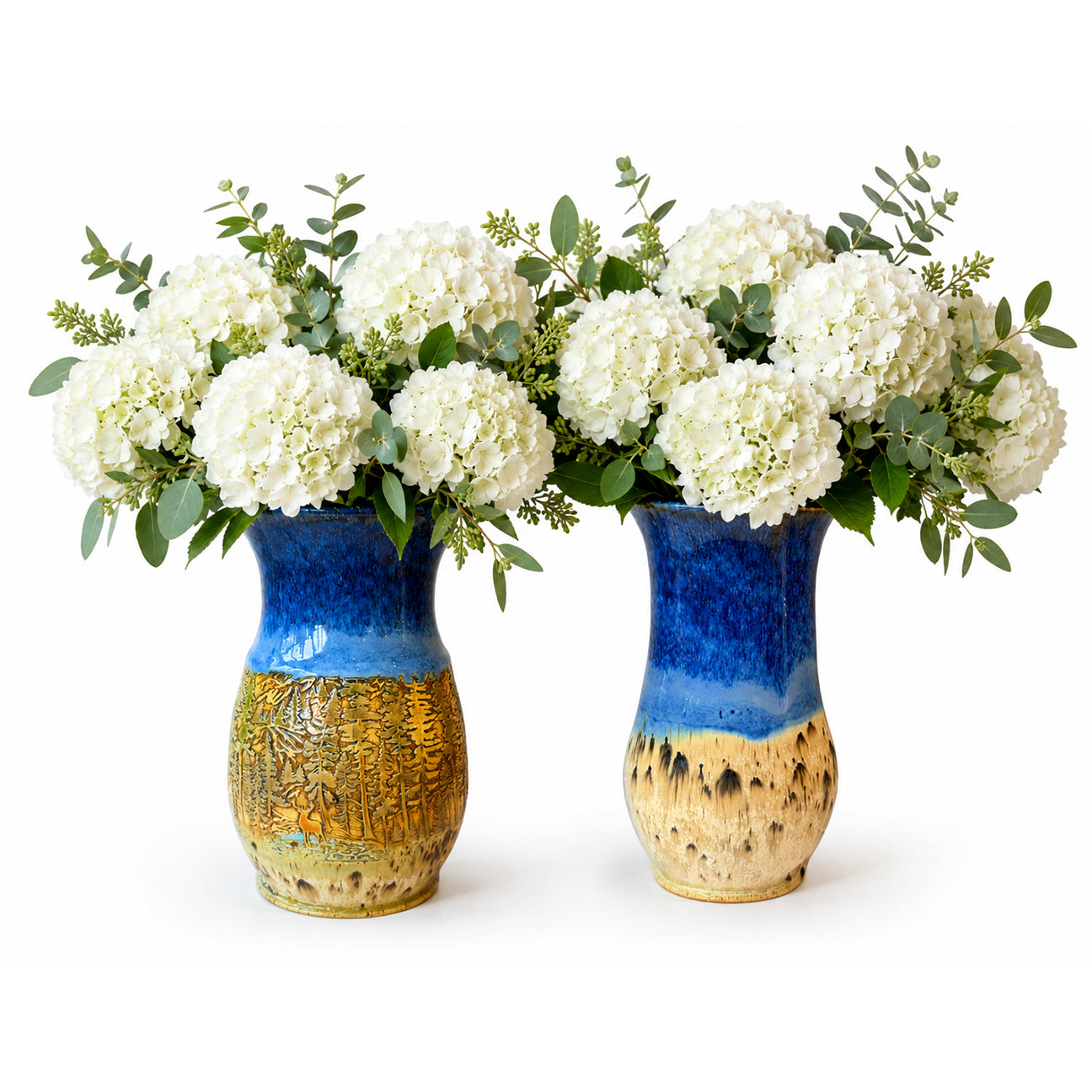 Two ceramic vases with a blue and gold glaze, filled with white hydrangea bouquets and green foliage, set against a plain white background.