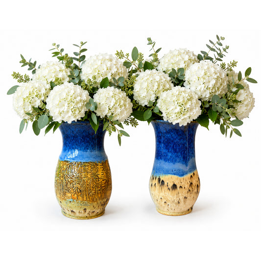 Two ceramic vases with a blue and gold glaze, filled with white hydrangea bouquets and green foliage, set against a plain white background.