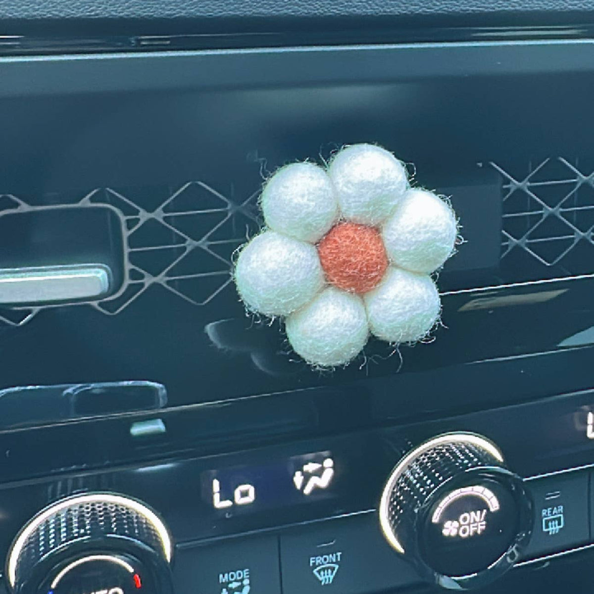 A small flower-shaped car air freshener with white fuzzy petals and a textured orange center, resting on a car's dashboard air vent.