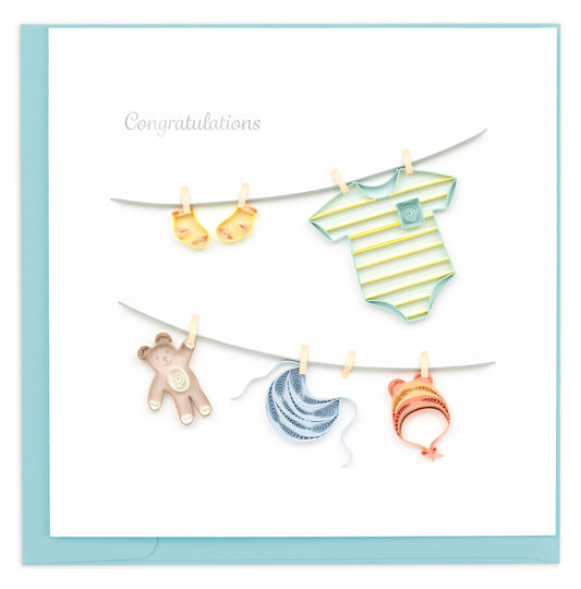 A greeting card featuring a handcrafted design with various baby clothes hanging on a clothesline, including a striped onesie, a teddy bear, and a pair of booties. The word 'Congratulations' is written at the top.